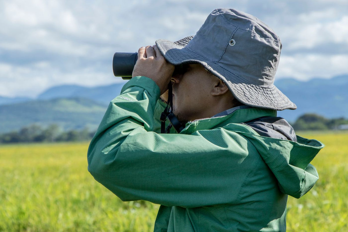 Someone in a green jacket and bucket hat looking through binoculars across a field
